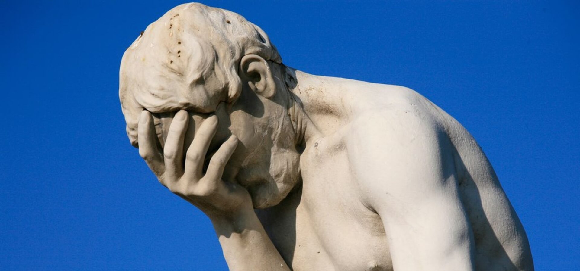 A stone statue covering its face with one hand, against a blue sky